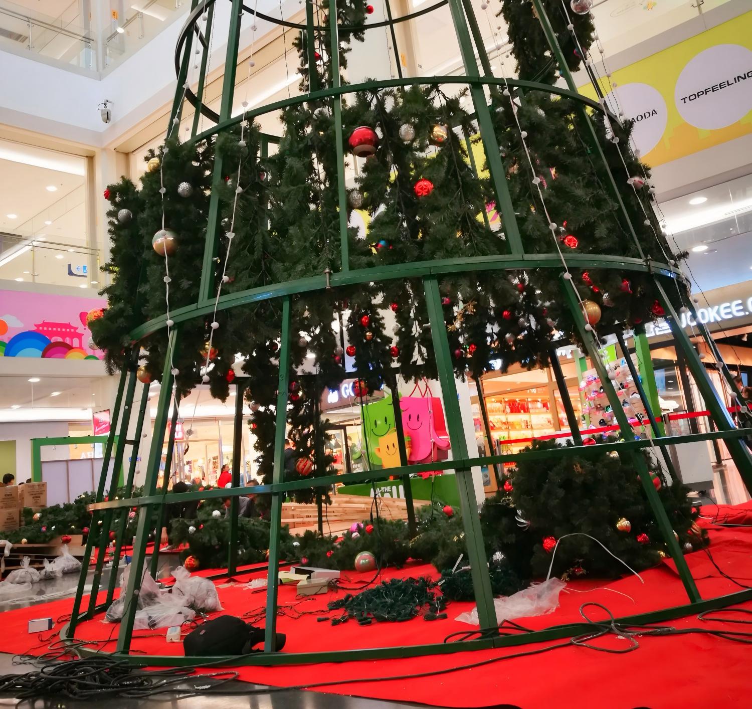 Large indoor commercial Christmas tree under installation with structural frame, artificial branches, ornaments, and visible wiring in a shopping mall