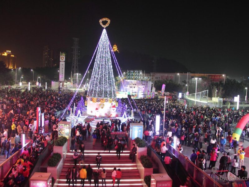 Large commercial Christmas tree in a plaza attracting crowds with supporting holiday decorations around the base