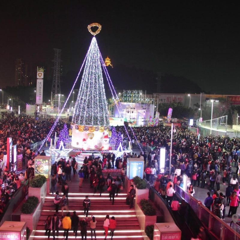Large commercial Christmas tree in a plaza attracting crowds with supporting holiday decorations around the base